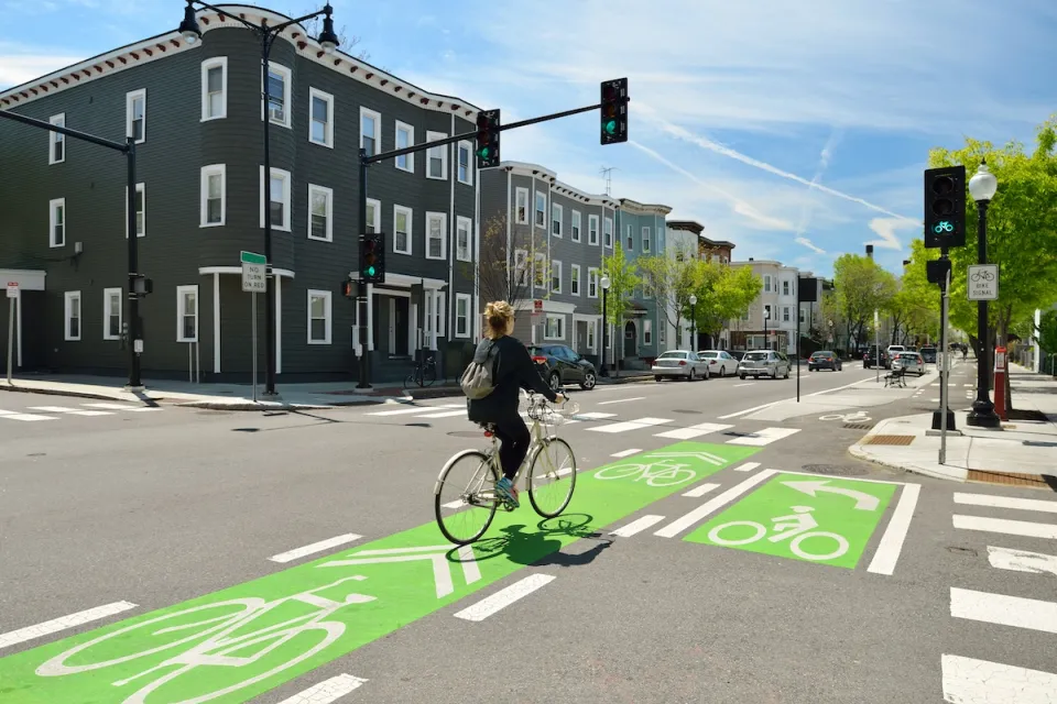 Cyclist on bike in green painted bike lane at intersection with three-story buildings in background.